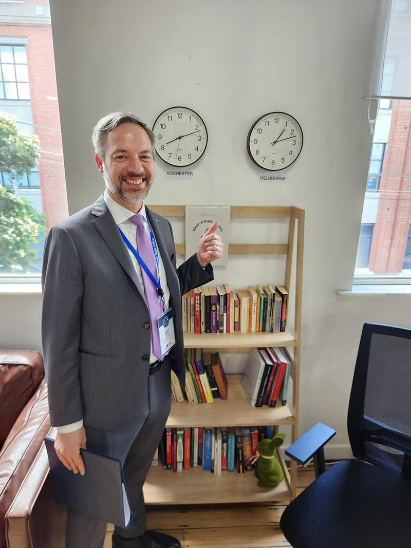 Commissioner Varilek points to two clocks on a wall, one with Melbourne's current time and one with Rochester's current time.