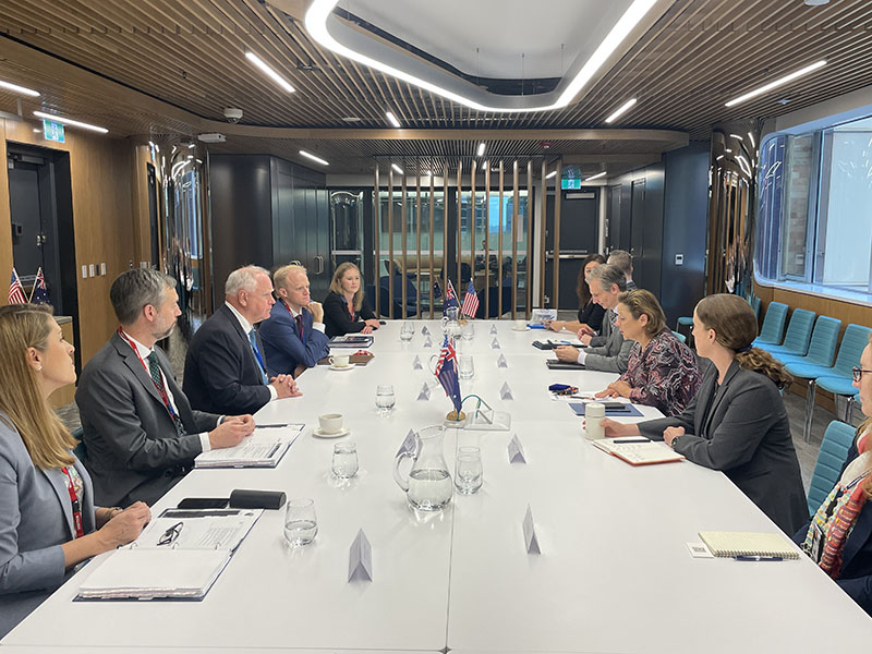 Multiple sit on two sides of a long table, facing each other and speaking.