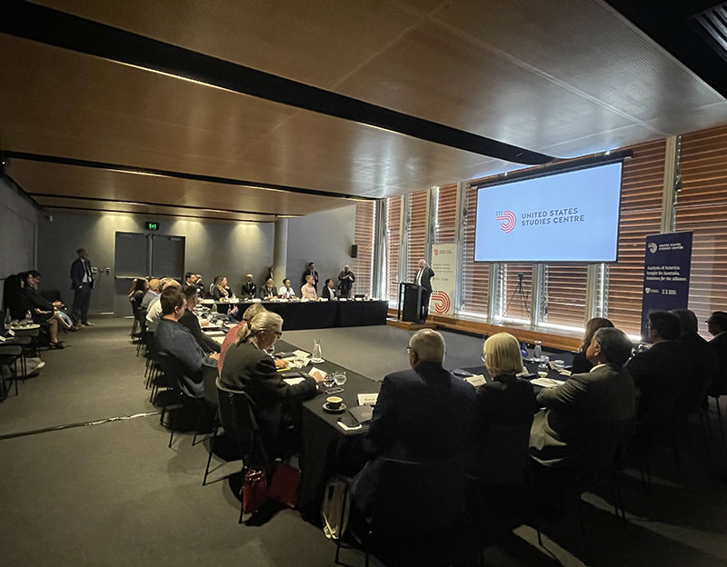 Picture of people sitting around a large table, listening to a speaker standing at a podium.